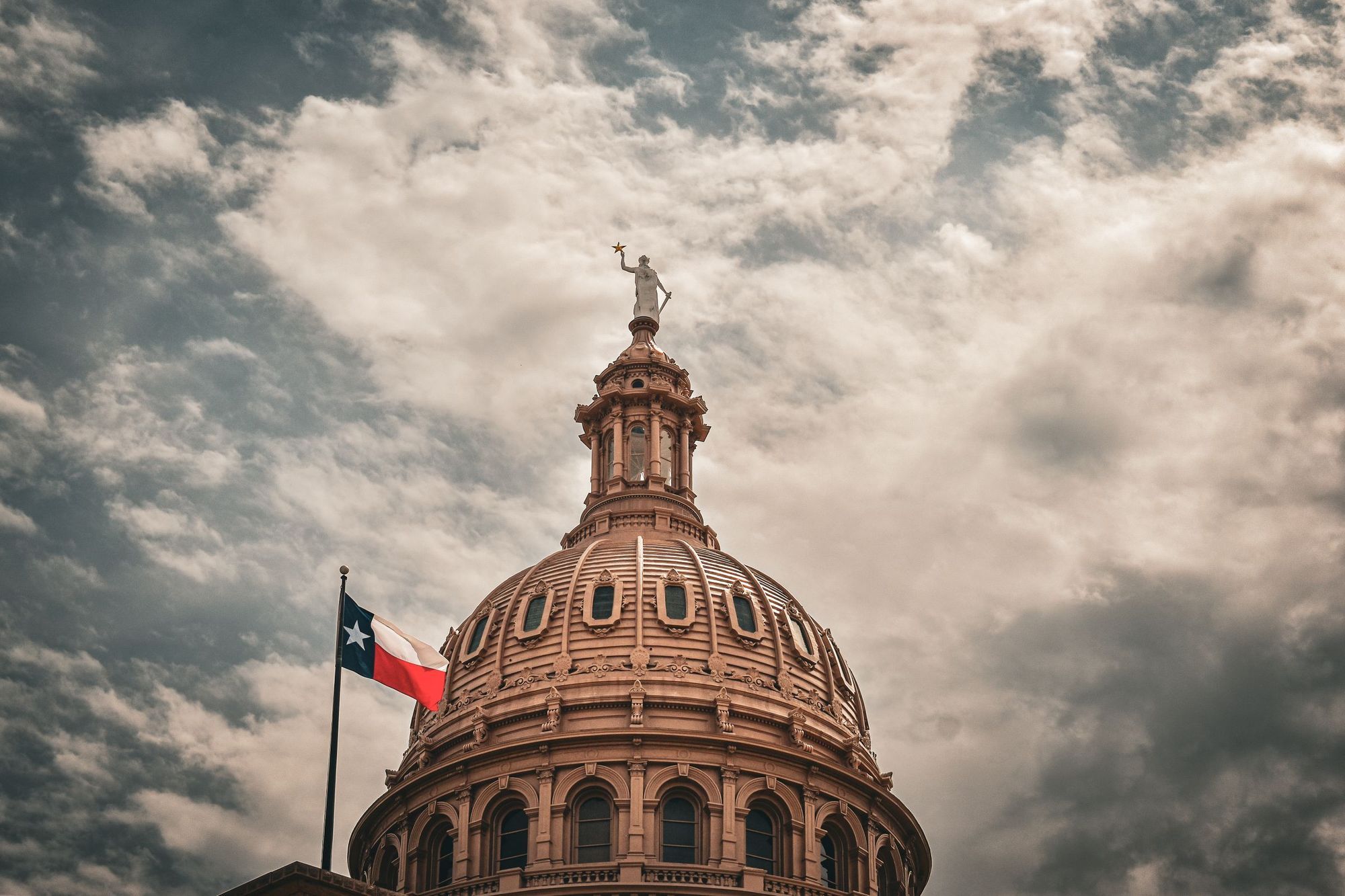 The dome of the Texas State Capitol with a Texas flag under a cloudy sky